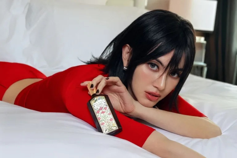 Modelka w czerwonej stylizacji prezentuje flakon perfum Gucci Flora na tle jasnego wnętrza. Perfumy należą do kluczowych kategorii Gucci Beauty, której rozwój i dystrybucję L’Oréal przejmie od Coty 1 lipca 2027 roku.