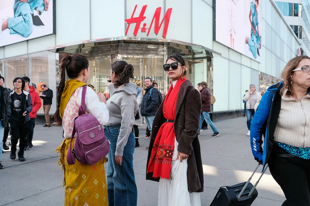 Wejście do sklepu H&M z czerwonym logo nad drzwiami, przed którym stoją i przechodzą klienci.