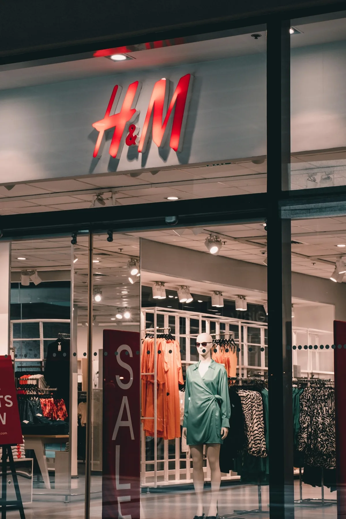 Witryna sklepu H&M z widocznym logo, manekinem, wieszakami z ubraniami i tablicą informującą o wyprzedaży.