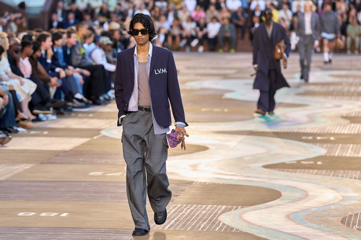 LVMH wyniki finansowe 2026. Model idzie po wybiegu podczas pokazu Louis Vuitton, prezentując stylizację z granatową marynarką, szarymi spodniami i niewielką torbą z monogramem marki. W tle widać publiczność oraz innych modeli. Zdjęcie nawiązuje do segmentu mody i galanterii skórzanej LVMH, który pozostaje kluczowym obszarem działalności grupy.