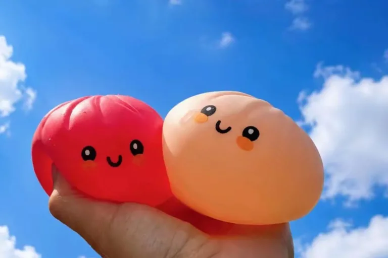 Dwa miękkie gniotki Mystery Squishy Dumplings w kolorze czerwonym i brzoskwiniowym są trzymane w dłoni na tle błękitnego nieba z chmurami. Obie zabawki mają uśmiechnięte buzie i kształt przypominający pierożki.