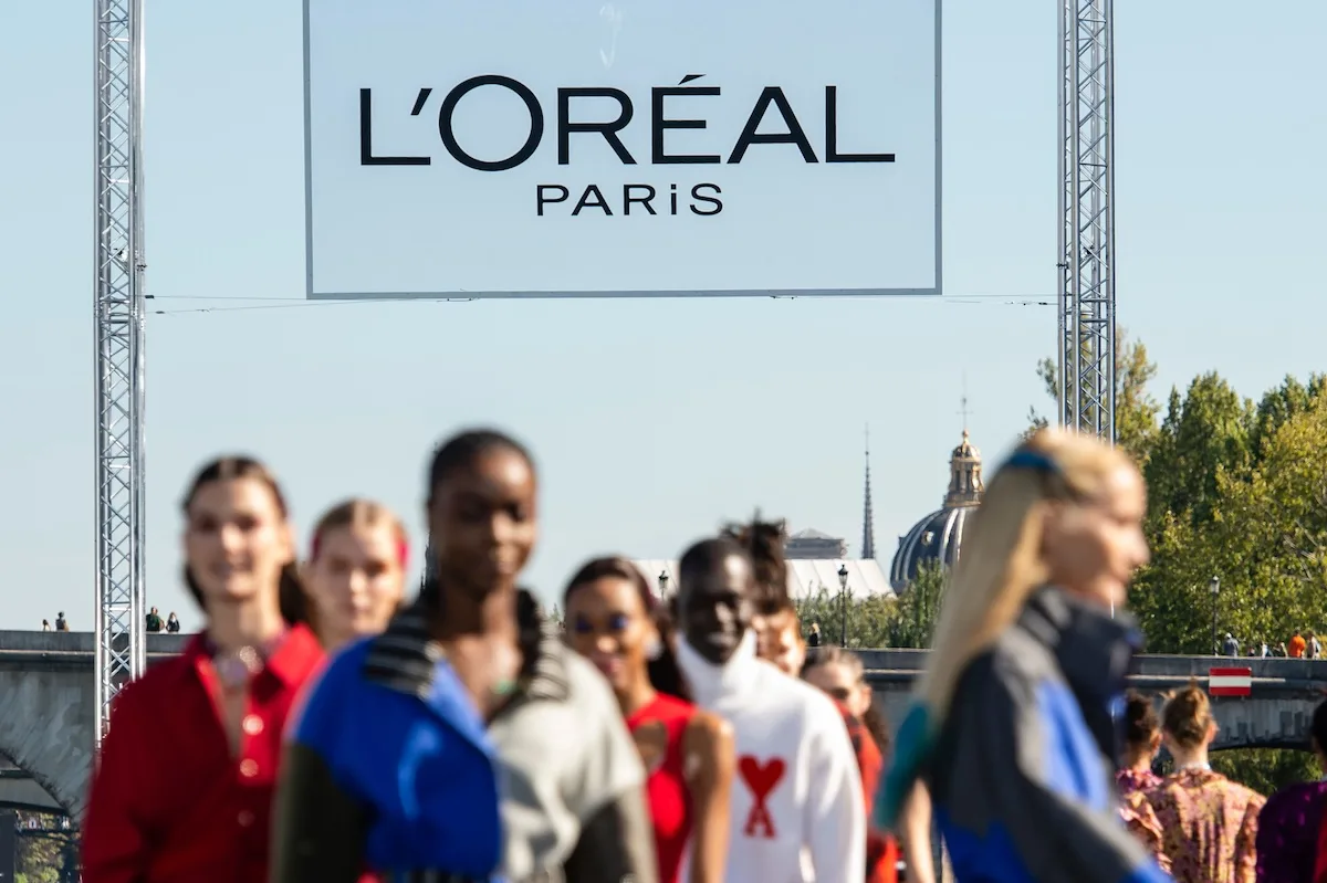 Rozmyte sylwetki modelek idących po wybiegu podczas pokazu L’Oréal Paris, nad którym góruje duże logo marki. Scena rozgrywa się w plenerze, na tle paryskiej architektury i błękitnego nieba. L’Oréal Paris zajęło pierwsze miejsce w rankingu najbardziej pożądanych pracodawców branży beauty 2026.