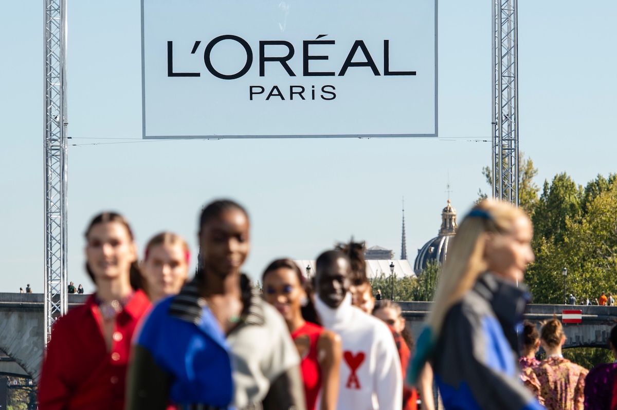 Rozmyte sylwetki modelek idących po wybiegu podczas pokazu L’Oréal Paris, nad którym góruje duże logo marki. Scena rozgrywa się w plenerze, na tle paryskiej architektury i błękitnego nieba. L’Oréal Paris zajęło pierwsze miejsce w rankingu najbardziej pożądanych pracodawców branży beauty 2026.