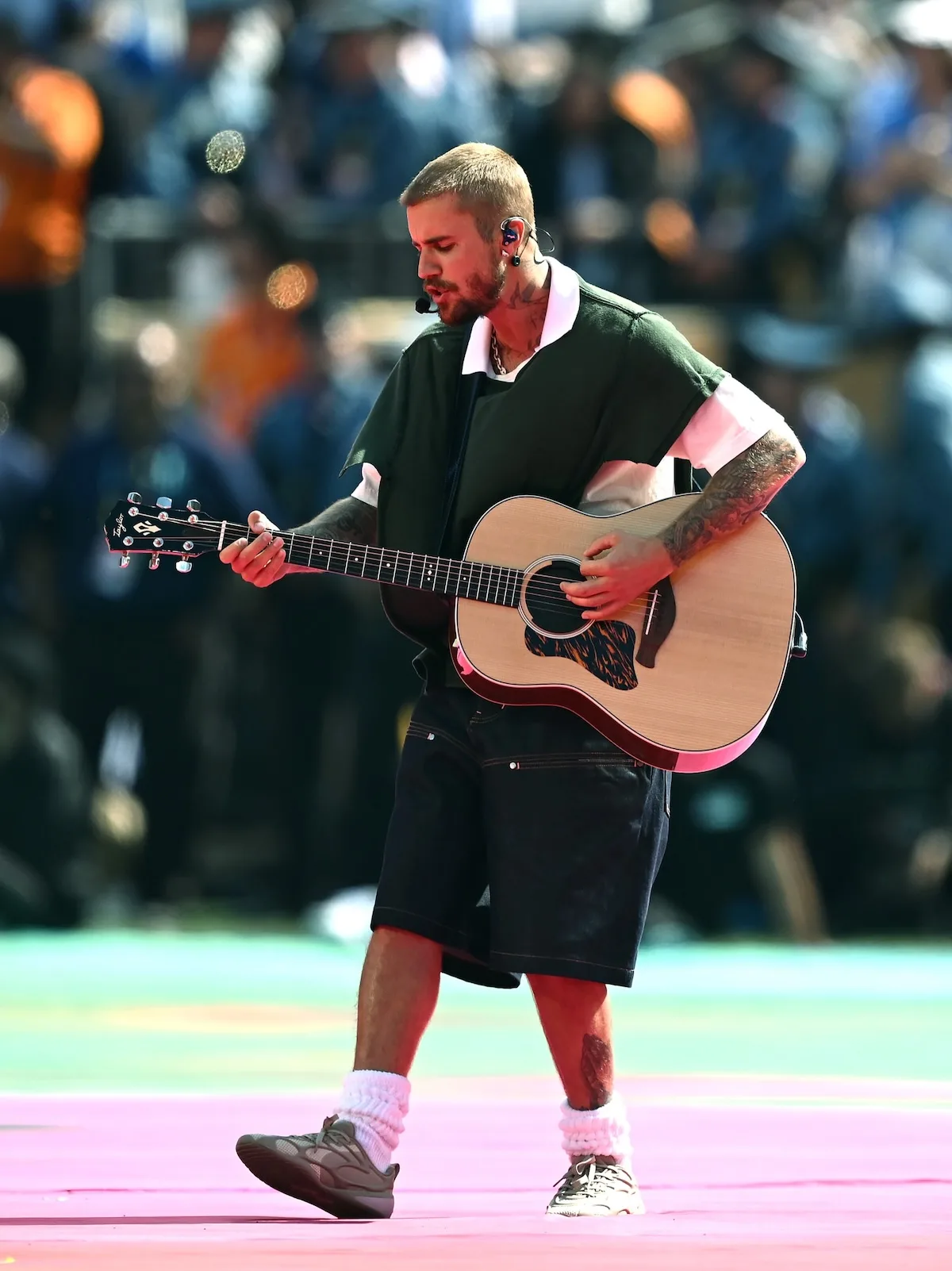 Justin Bieber gra na gitarze akustycznej podczas halftime show finału mistrzostw świata 2026. Występuje w czarno-białym polo, szerokich dżinsowych szortach, grubych skarpetach i sportowych sneakersach, prezentując swobodną estetykę swojej marki Skylrk.