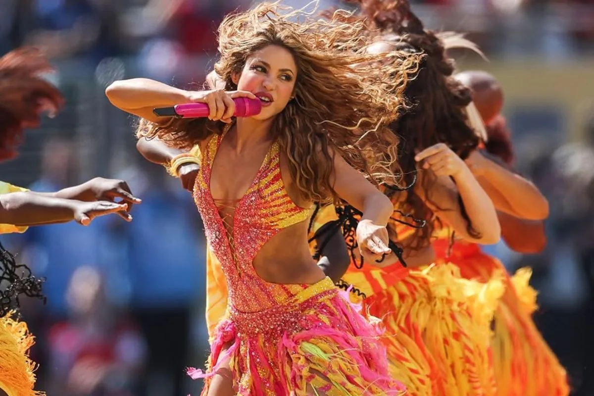 Shakira śpiewa do różowego mikrofonu podczas halftime show finału mistrzostw świata 2026. Występuje w bogato zdobionym kostiumie z kryształami i kolorowymi frędzlami, a dynamiczną choreografię uzupełniają tancerze w ognistych stylizacjach.