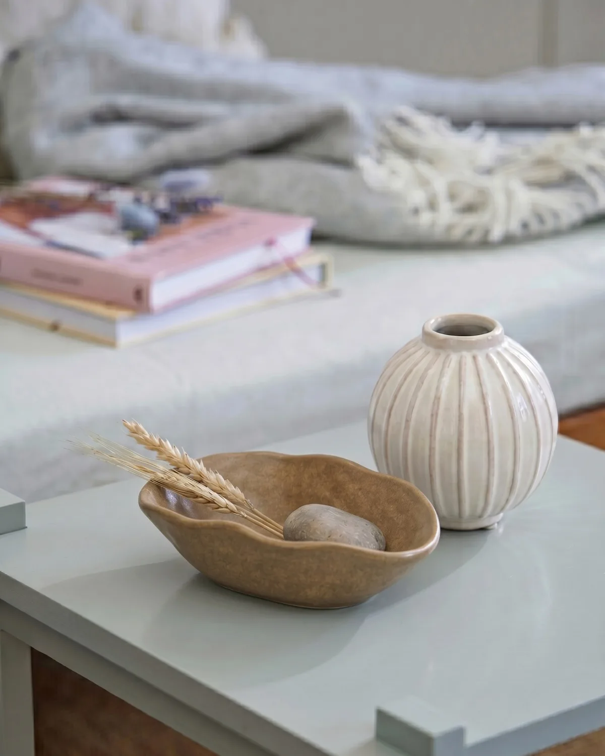 Zdjęcie przedstawia stolik kawowy z dekoracyjną ceramiką Søstrene Grene – jasnym wazonem, nieregularną misą i naturalnymi dodatkami. W tle widać miękkie tekstylia i książki, które podkreślają spokojny, skandynawski charakter aranżacji.