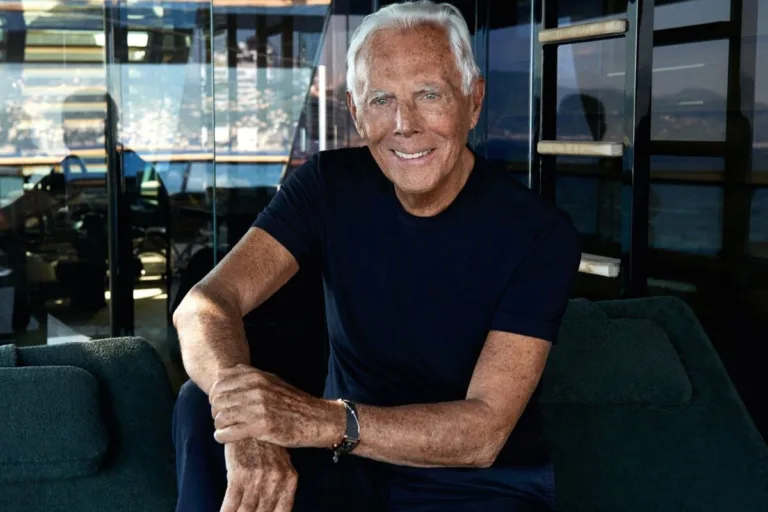 Serial o Giorgio Armanim. Giorgio Armani siedzi na ciemnej sofie w nowoczesnym wnętrzu z dużymi, przeszklonymi ścianami. Projektant ma na sobie prosty granatowy T-shirt i spodnie w podobnym kolorze, a na nadgarstku nosi zegarek. Uśmiecha się do obiektywu, a w tle widać odbijające się w szybach światło oraz fragment nadmorskiego krajobrazu.
