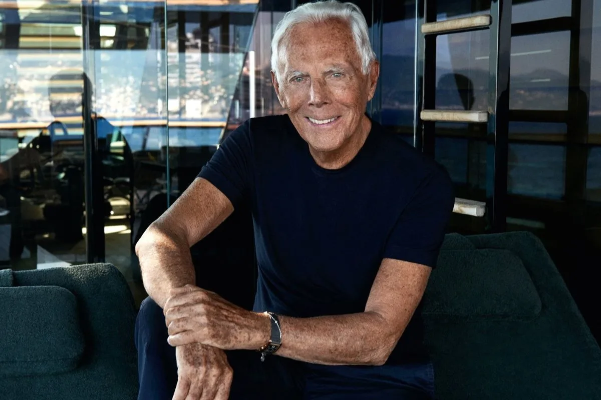 Serial o Giorgio Armanim. Giorgio Armani siedzi na ciemnej sofie w nowoczesnym wnętrzu z dużymi, przeszklonymi ścianami. Projektant ma na sobie prosty granatowy T-shirt i spodnie w podobnym kolorze, a na nadgarstku nosi zegarek. Uśmiecha się do obiektywu, a w tle widać odbijające się w szybach światło oraz fragment nadmorskiego krajobrazu.