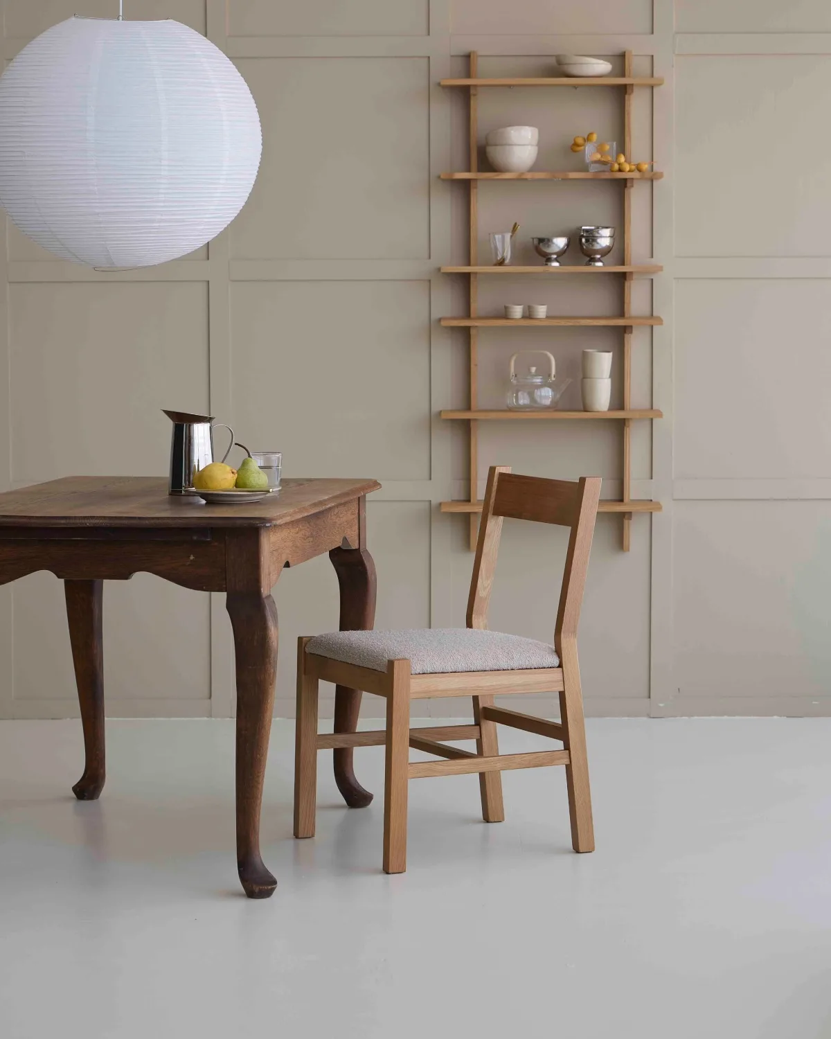 Aranżacja jadalni w skandynawskim stylu z drewnianym stołem, krzesłem, papierową lampą i ścienną półką z ceramiką. Jasne kolory, naturalne materiały i proste dodatki nawiązują do estetyki marki Søstrene Grene.
