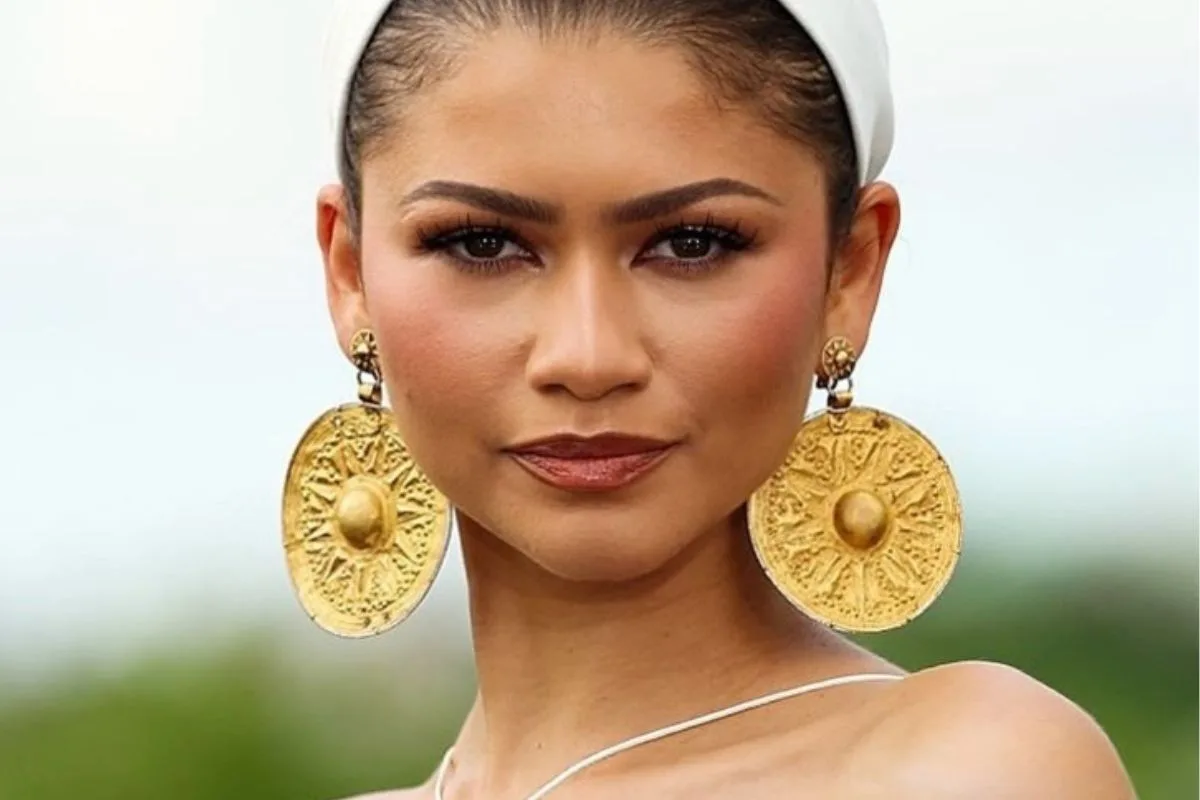 Zendaya kolczyki. Zendaya pozuje w białej opasce i minimalistycznej sukni na cienkich ramiączkach. Po obu stronach jej twarzy widoczne są duże, okrągłe złote kolczyki ozdobione promienistym motywem. Kontrowersyjna biżuteria stanowi centralny element całej stylizacji aktorki.