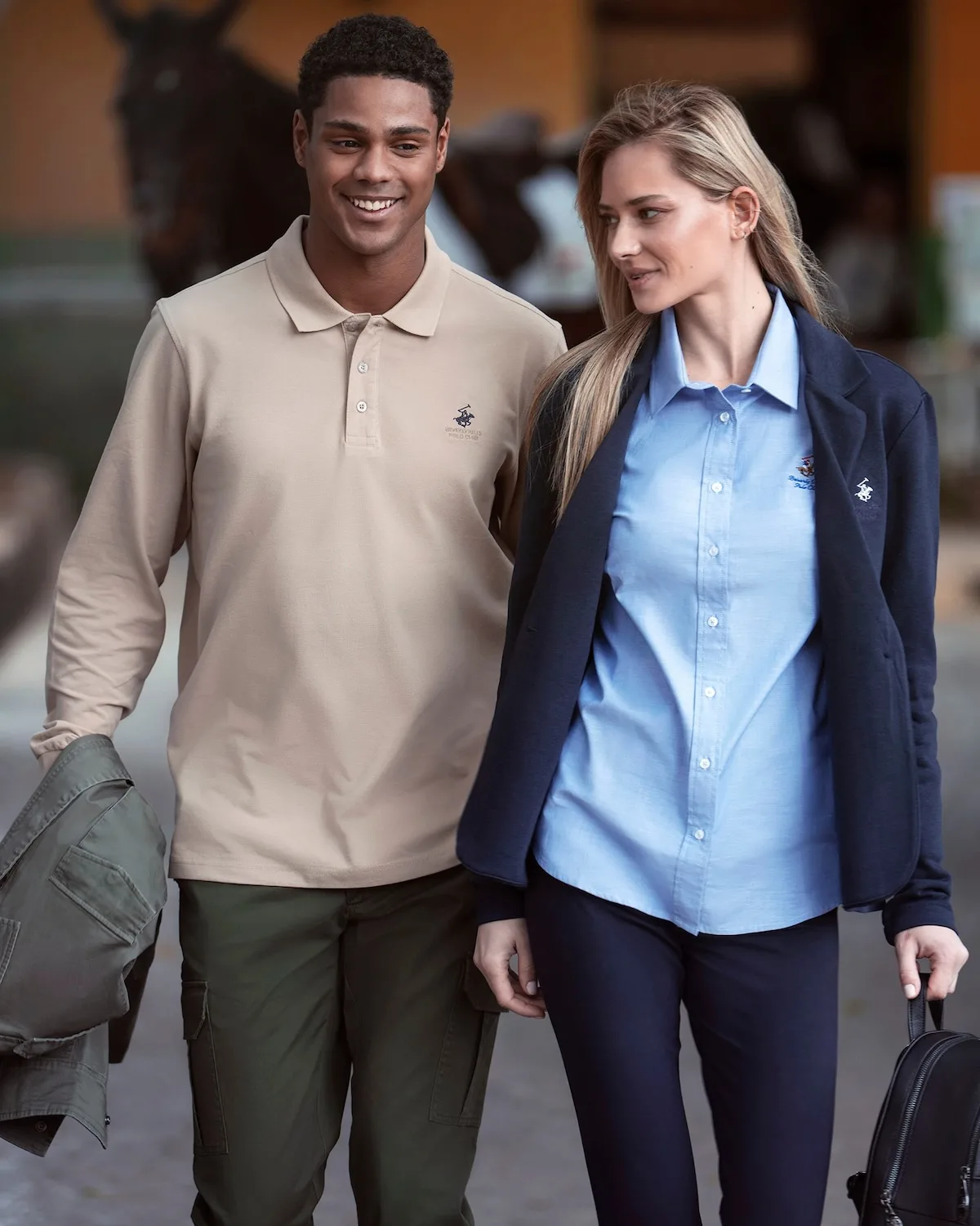 Model i modelka prezentują stylizacje Beverly Hills Polo Club w eleganckim, casualowym wydaniu. Mężczyzna ma na sobie beżową koszulkę polo z logo marki, a kobieta błękitną koszulę i granatową marynarkę. Zdjęcie pokazuje klasyczną, codzienną ofertę odzieżową BHPC inspirowaną stylem preppy.