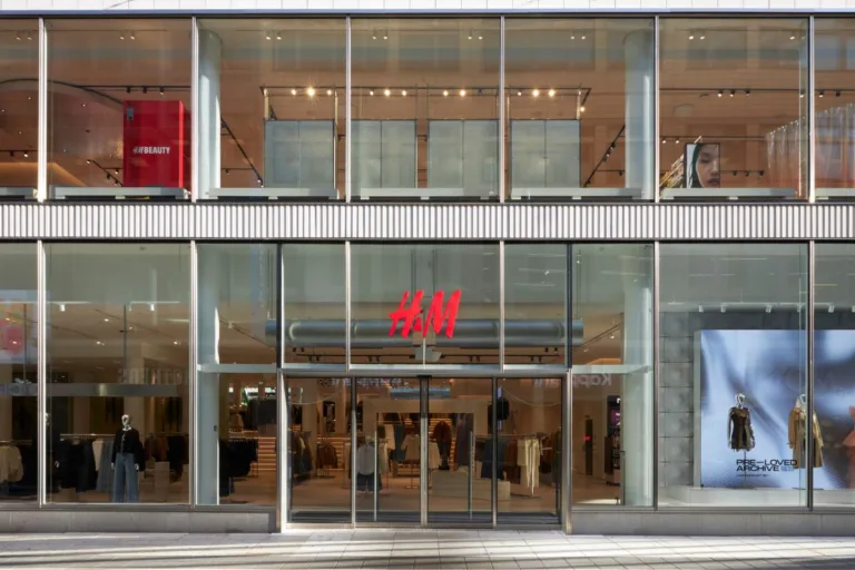 H&M zwolnienia 2026. Fasada sklepu H&M z dużymi przeszklonymi witrynami i czerwonym logo nad wejściem. W witrynach widoczne są manekiny, ekspozycja odzieży oraz strefa H&M Beauty na piętrze.