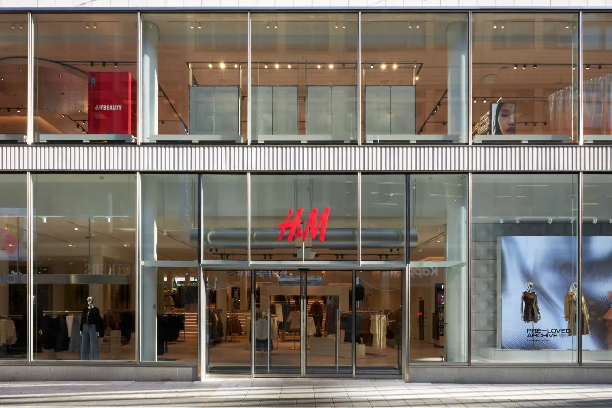 H&M zwolnienia 2026. Fasada sklepu H&M z dużymi przeszklonymi witrynami i czerwonym logo nad wejściem. W witrynach widoczne są manekiny, ekspozycja odzieży oraz strefa H&M Beauty na piętrze.