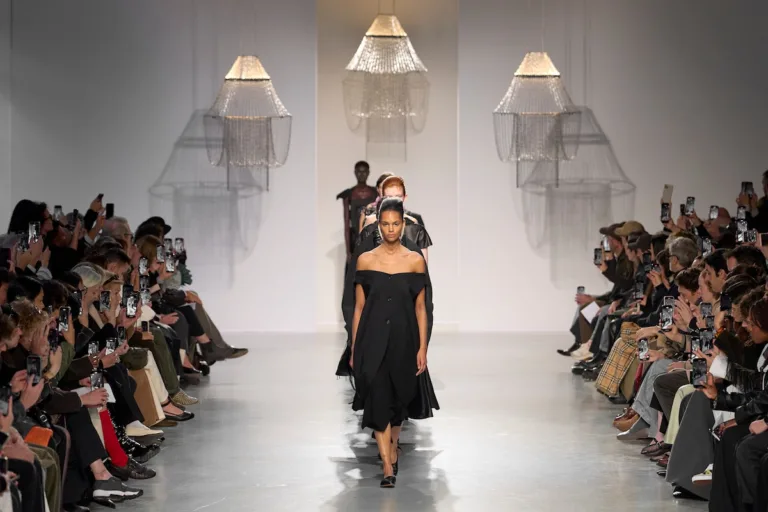 Harmonogram Fashion Week SS2027. Modelki prezentują kolekcję Pasqualetti podczas pokazu na minimalistycznym wybiegu. Na pierwszym planie widać czarną sukienkę z odkrytymi ramionami, a po obu stronach publiczność fotografującą finał prezentacji.
