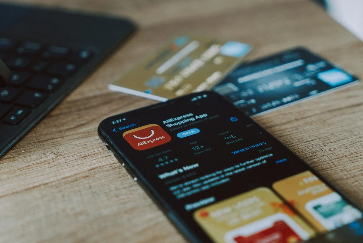 AliExpress: Smartfon leży na drewnianym biurku z otwartą stroną aplikacji AliExpress Shopping App w sklepie z aplikacjami. W tle znajdują się dwie karty płatnicze oraz fragment laptopa.