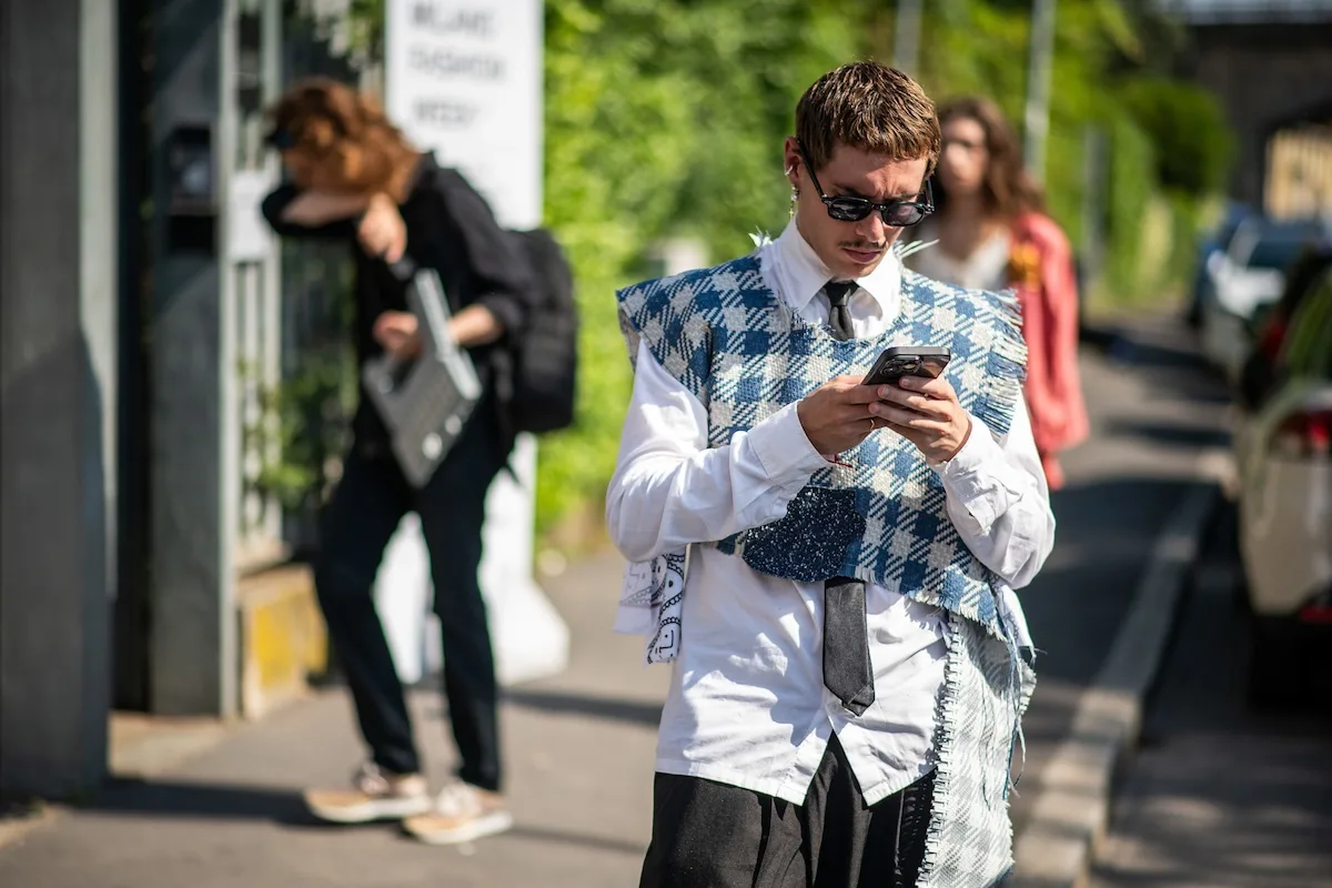 Temu i AliExpress tracą klientów przez cło na przesyłki z Chin. Mężczyzna w białej koszuli, czarnym krawacie i niebiesko-białej kraciastej kamizelce korzysta ze smartfona na ulicy. Ma na sobie ciemne okulary, a w tle widać przechodniów, samochód i zieleń.