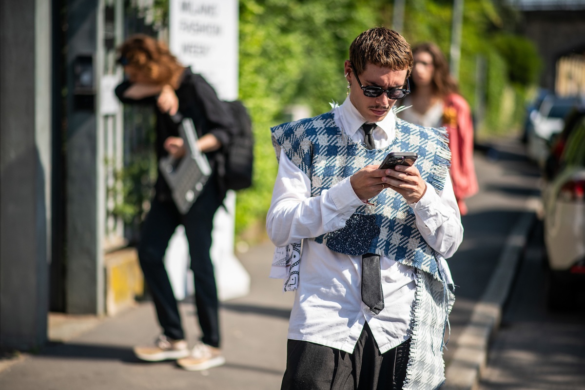 Temu i AliExpress tracą klientów przez cło na przesyłki z Chin. Mężczyzna w białej koszuli, czarnym krawacie i niebiesko-białej kraciastej kamizelce korzysta ze smartfona na ulicy. Ma na sobie ciemne okulary, a w tle widać przechodniów, samochód i zieleń.