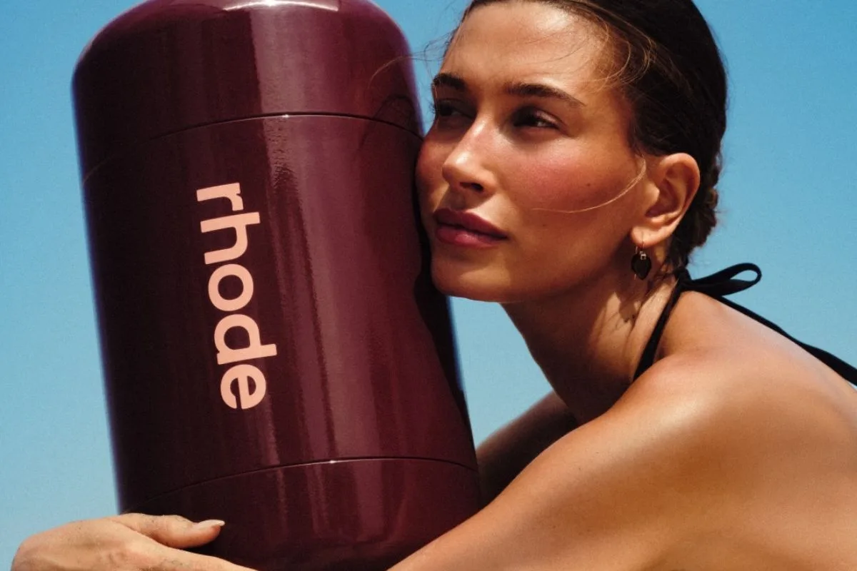 Sprzedaż Rhode wyniki. Hailey Bieber w kampanii reklamowej Rhode obejmuje duże brązowe opakowanie z wyraźnym logo marki. W tle znajduje się intensywnie błękitne niebo, a fotografia promuje letnią kolekcję kosmetyków Rhode.