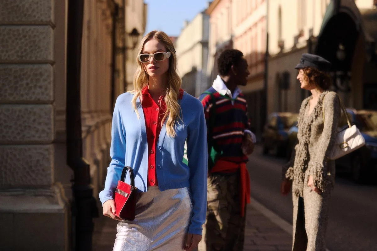 Kobieta w błękitnym kardiganie i czerwonej koszuli pozuje na ulicy w otoczeniu modnie ubranych osób. Stylizacja nawiązuje do estetyki street style znanej z tygodni mody.