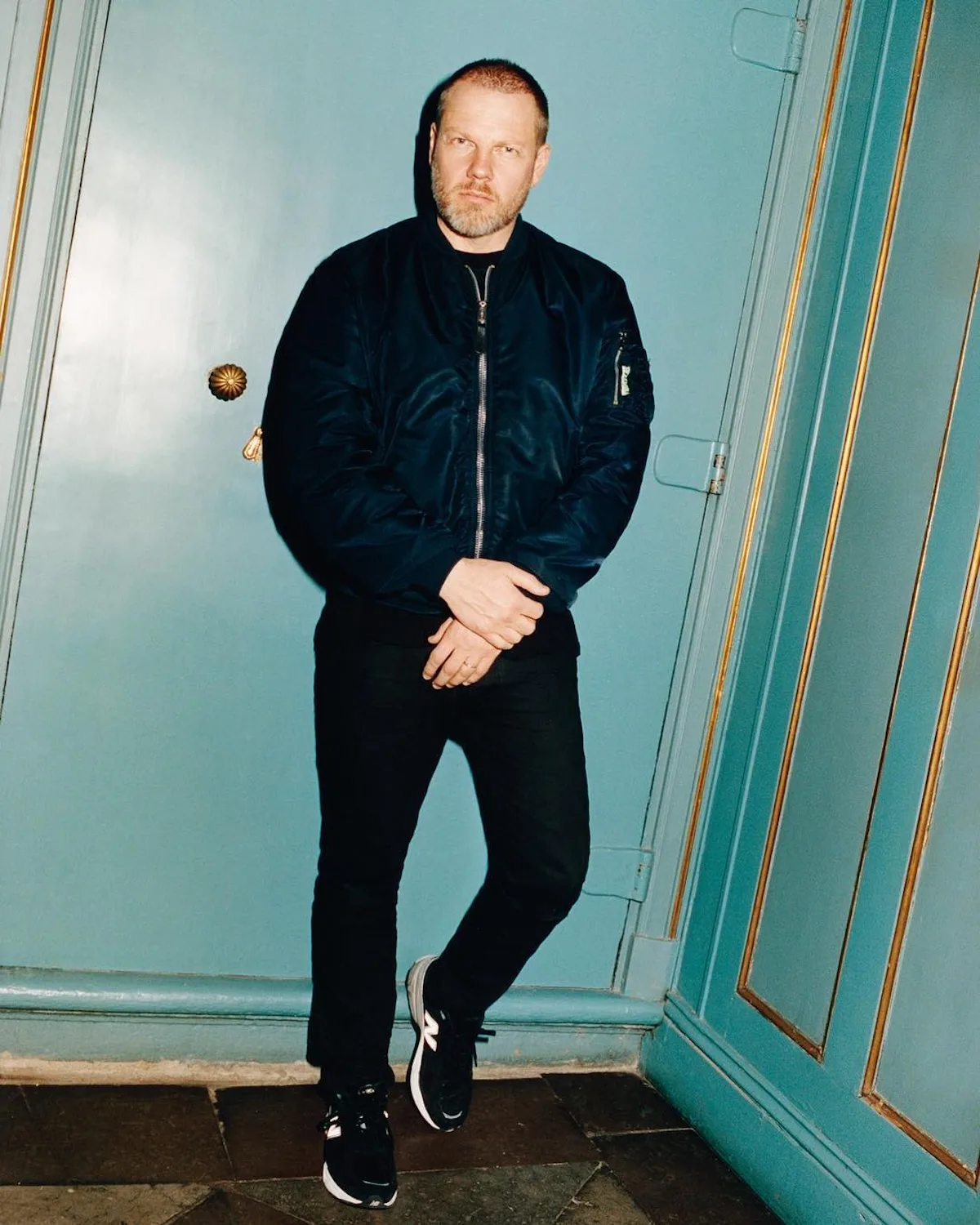 Jonny Johansson, współzałożyciel i dyrektor kreatywny Acne Studios, pozuje w jasnoniebieskim wnętrzu ze złotymi detalami. Ma na sobie czarną kurtkę bomber, czarne spodnie i sportowe buty.