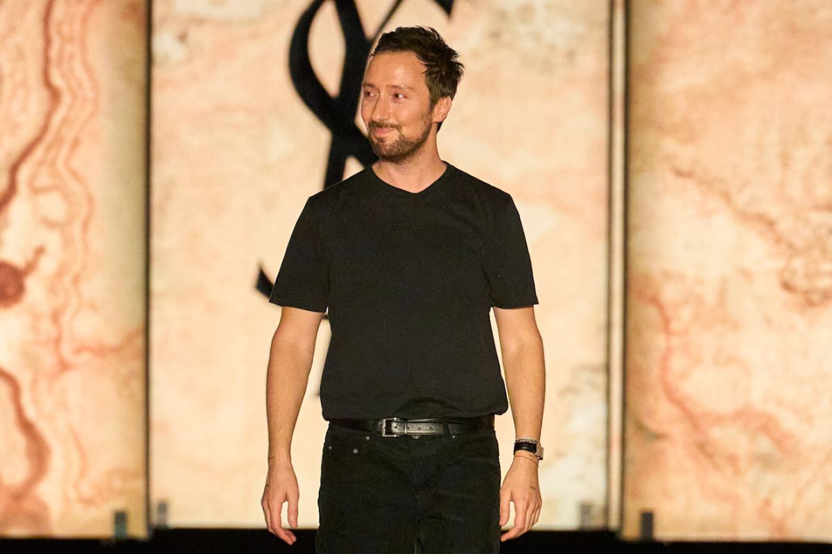 Anthony Vaccarello odchodzi z Saint Laurent? Na zdjęciu Anthony Vaccarello podczas wyjścia na wybieg po pokazie mody. Projektant stoi na scenie w czarnym T-shircie i czarnych spodniach, lekko się uśmiechając i spoglądając w bok. W tle widoczne są podświetlone dekoracje oraz charakterystyczne logo Saint Laurent.
