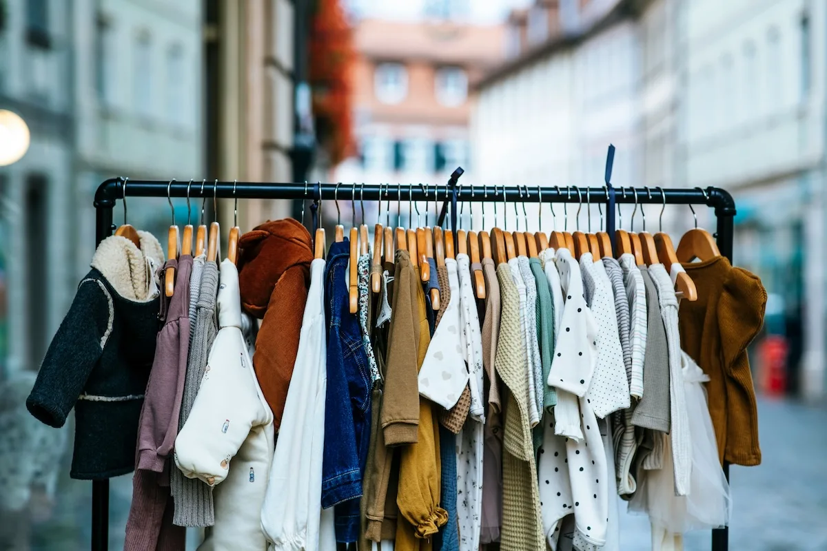 UOKiK sprawdził składy ubrań dla dzieci. Wieszak z odzieżą dziecięcą ustawiony przy witrynie sklepowej. Na drewnianych wieszakach wiszą m.in. kurtki, swetry, spodnie i ubranka w jasnych, stonowanych kolorach.