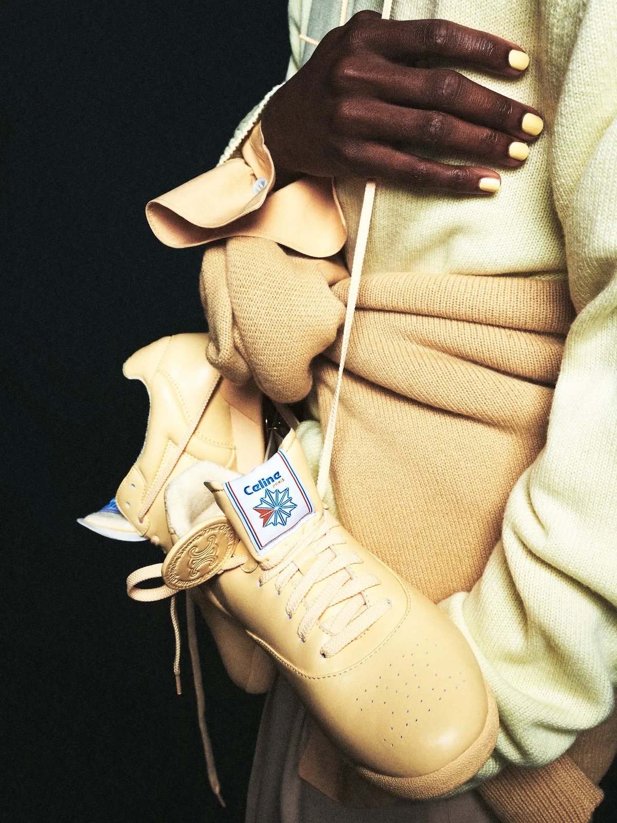Zbliżenie na kremowe sneakersy Celine x Reebok Freestyle Lo z jasnymi sznurowadłami i logo na języku, zestawione z beżową dzianiną i żółtym manicure.