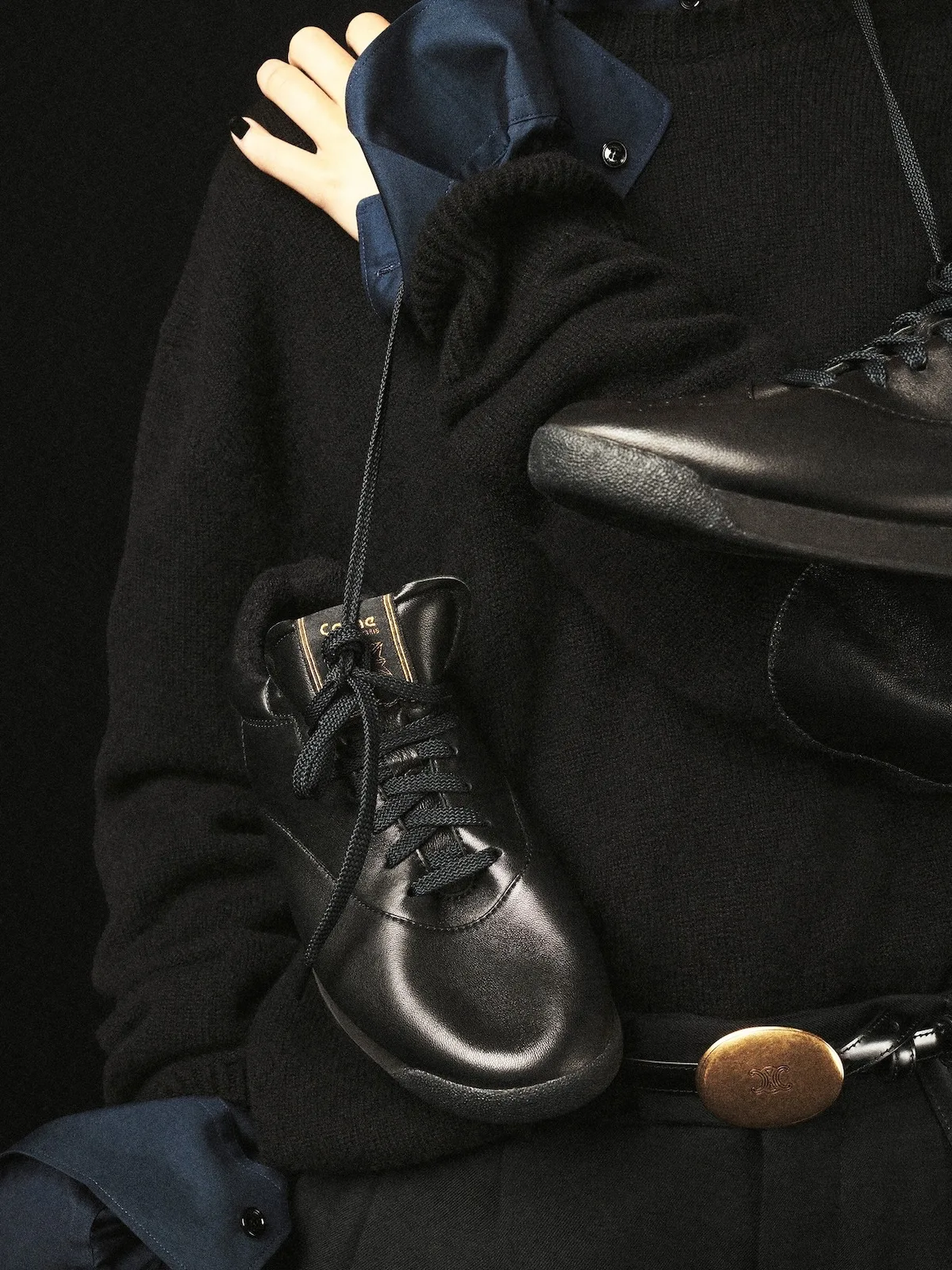 Zbliżenie na czarne sneakersy Celine x Reebok Freestyle Lo ze skórzaną cholewką, czarnymi sznurowadłami i logo marki, na tle ciemnej stylizacji z dzianiny.