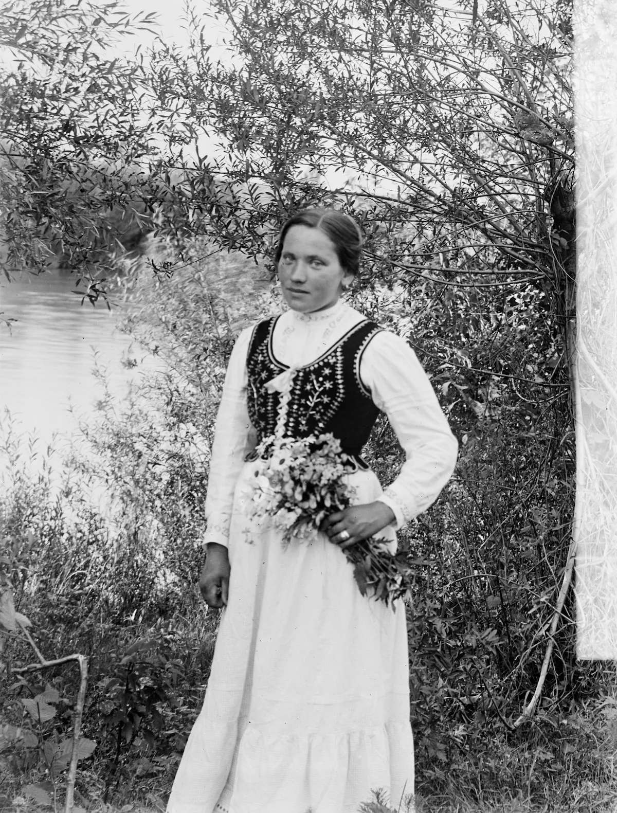 Archiwalna czarno-biała fotografia kobiety w tradycyjnym stroju ludowym, stojącej w ogrodzie z bukietem kwiatów. Jej ciemny gorset zdobią jasne hafty kwiatowe.