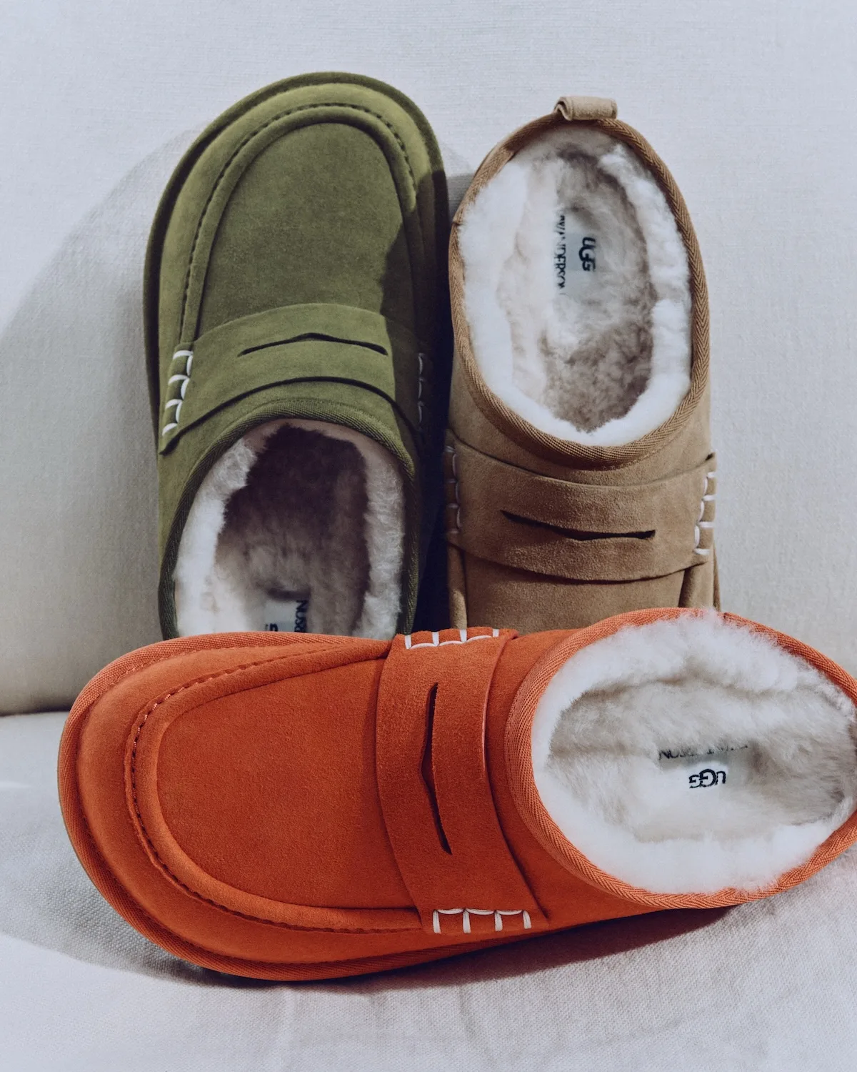 Kapcie UGG x JW Anderson sfotografowane z góry w kolorach khaki, beżowym i pomarańczowym, z miękkim futerkiem oraz paskami i przeszyciami jak w mokasynach.