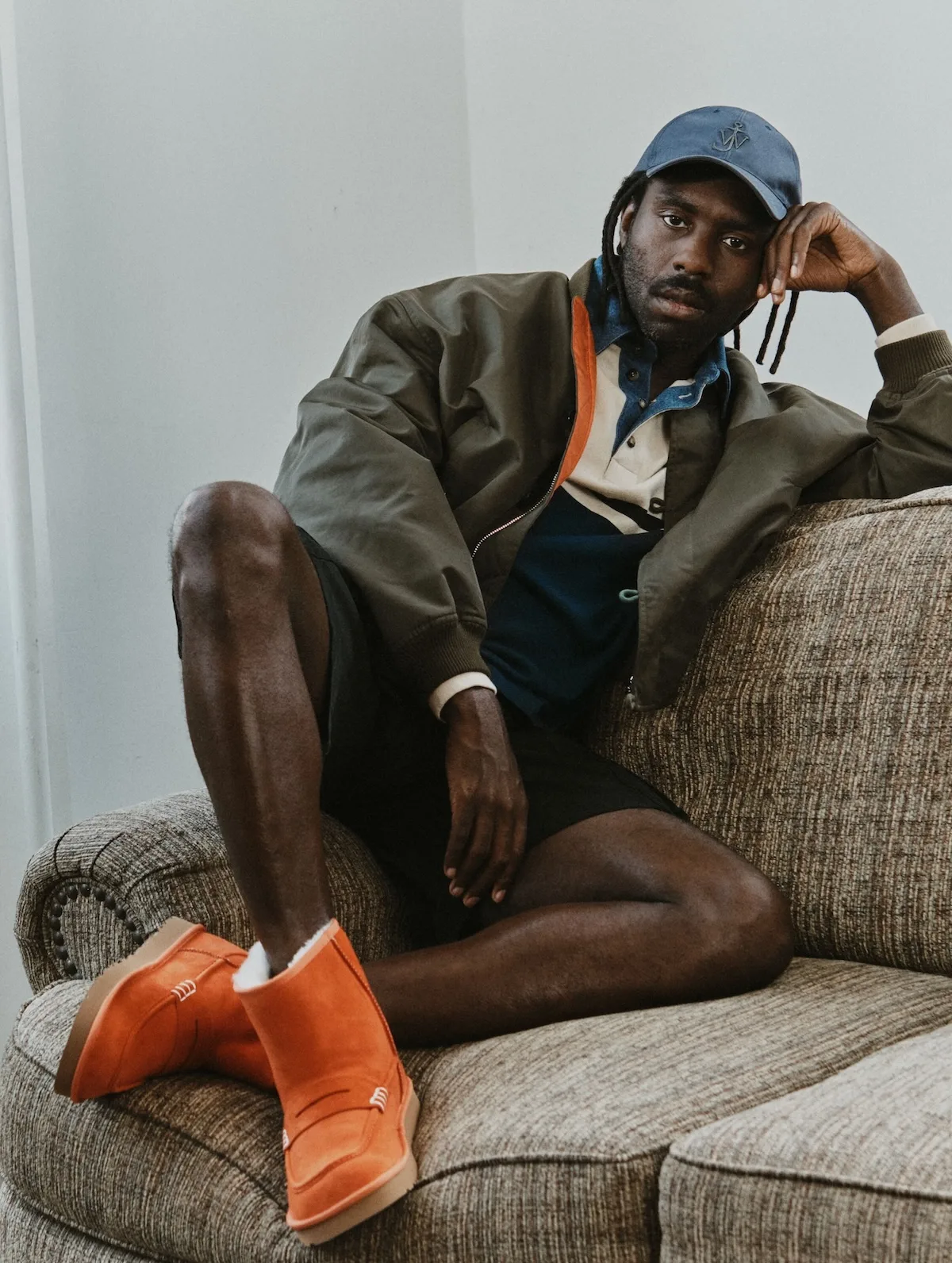 Dev Hynes pozuje na kanapie w oliwkowej kurtce, szortach i niebieskiej czapce, prezentując pomarańczowe mokasyny UGG x JW Anderson z ozdobnymi przeszyciami.