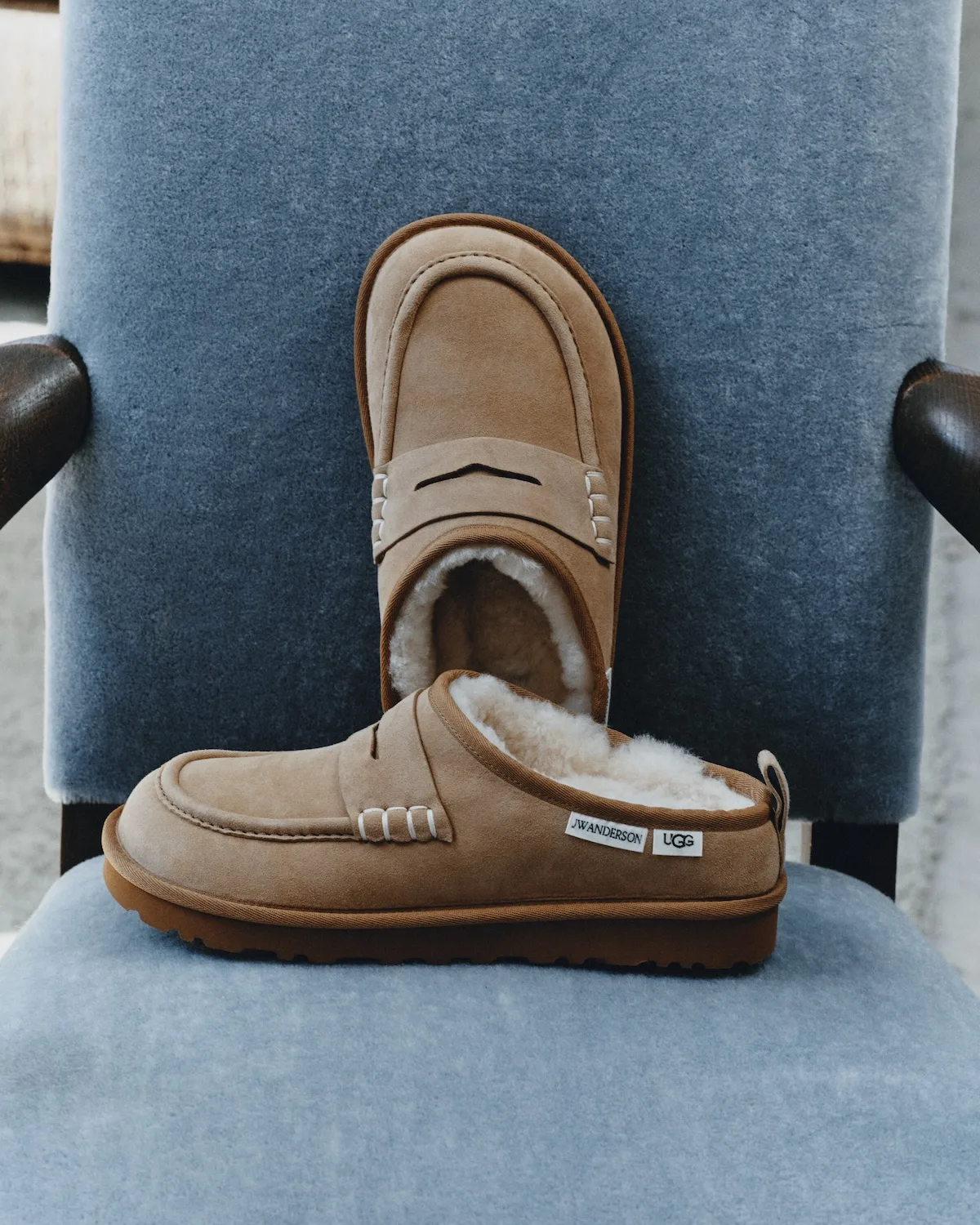 Beżowe mokasyny UGG x JW Anderson Loafer z zamszu, futrzanym wnętrzem i ozdobnymi przeszyciami, pokazane z góry i z boku na niebieskim tapicerowanym fotelu.