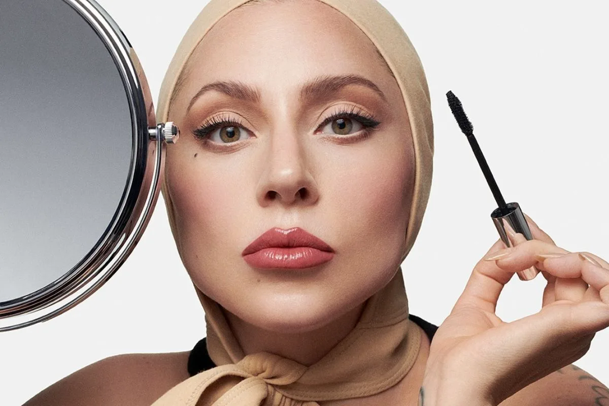 Lady Gaga CEO Haus Labs. Na zdjęciu Lady Gaga w kampanii Haus Labs, z delikatnym makijażem i beżowym nakryciem głowy, trzyma szczoteczkę do tuszu do rzęs obok okrągłego lusterka.