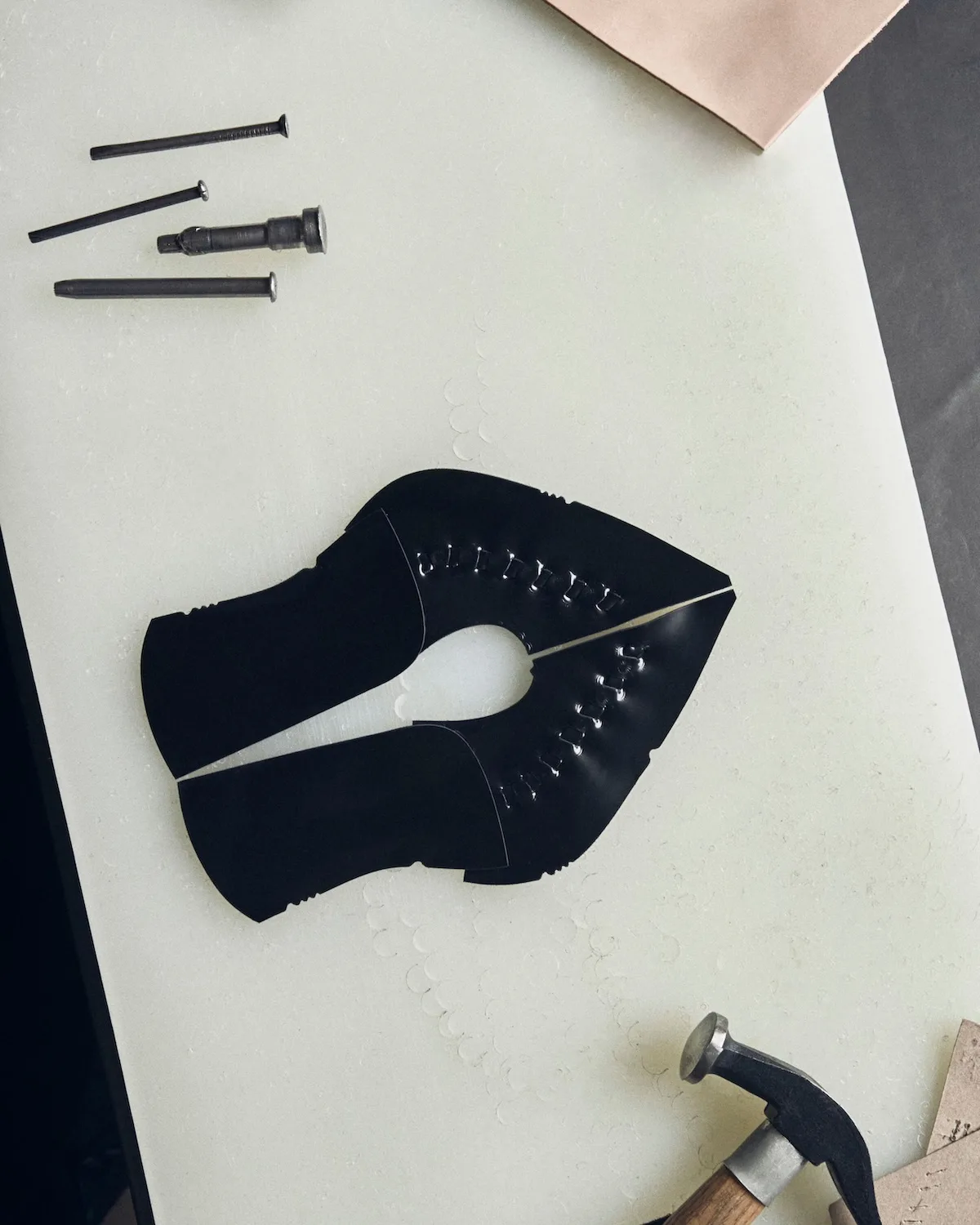 Czarne skórzane elementy obuwia z dekoracyjnymi przeszyciami, ułożone na jasnym blacie obok metalowych gwoździ i młotka. Zdjęcie z kampanii Podhale Leather Magdy Butrym, nawiązujące do procesu rzemieślniczego.