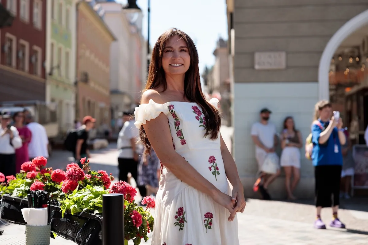 Dominika Kulczyk w białej sukience z odkrytymi ramionami i kwiatowym haftem, pozująca na tle kolorowych kamienic i różowych kwiatów.