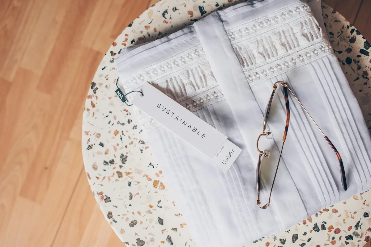Złożone białe ubranie z ozdobnymi haftami, metka z napisem „Sustainable Luxury” i okulary przeciwsłoneczne ułożone na stoliku z lastryko w ciepłym świetle dnia.