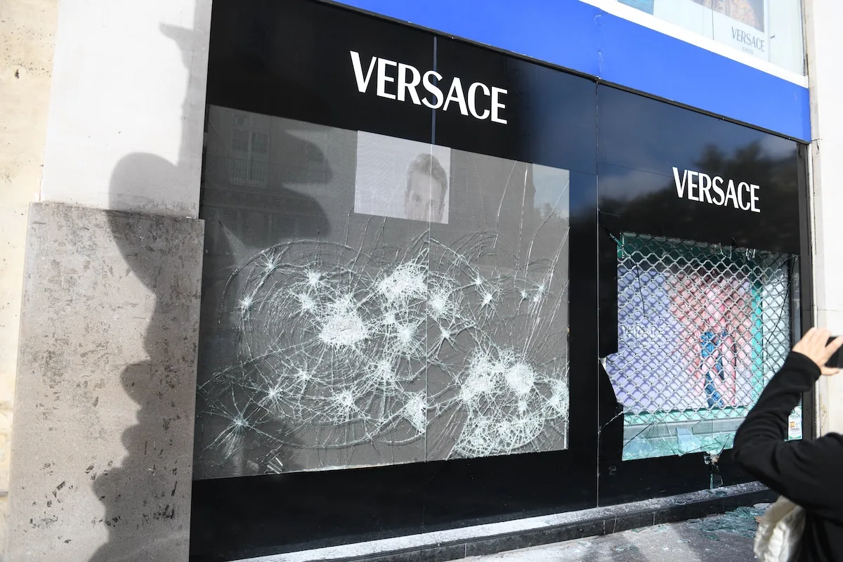 Rozbita witryna butiku Versace z widocznym logo luksusowej marki. Na szybach widać liczne ślady uderzeń, a przed sklepem leżą odłamki potłuczonego szkła.